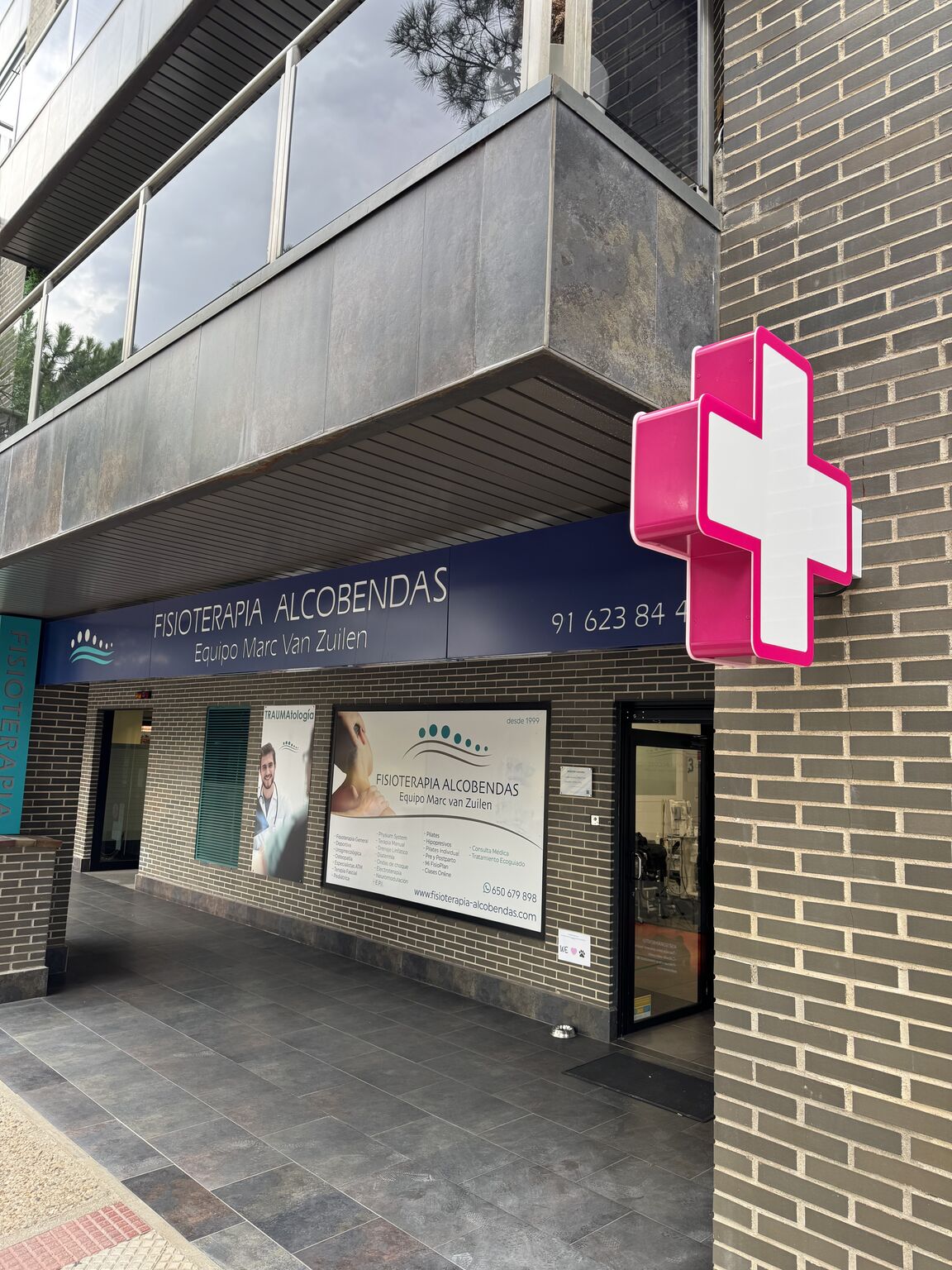 Fachada de Fisioterapia Alcobendas — Equipo Marc van Zuilen con el Símbolo Oficial de la Fisioterapia (cruz rosa magenta) instalado